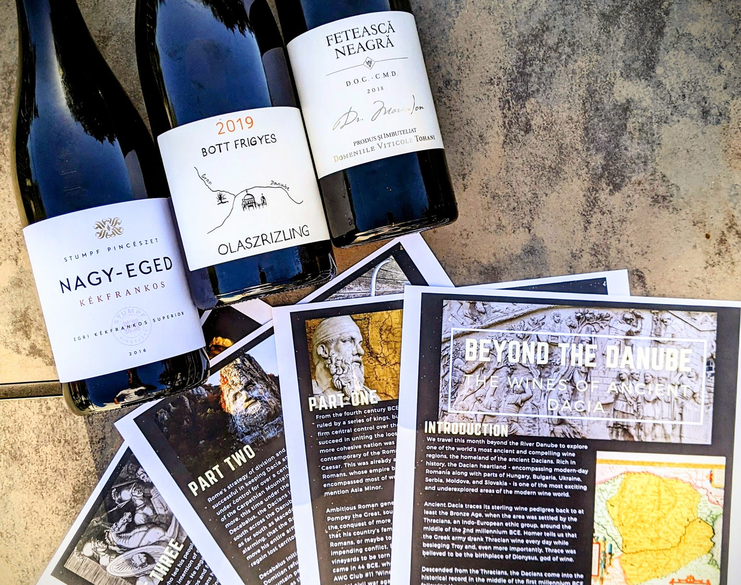 Three bottles of eastern european wine are laid out on a table next to four full color pages of ancient history plus wine essays by the Ancient Wine Guys