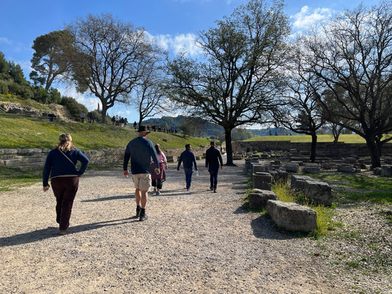 A group of five travelers on the Ancient Wine Guys trip to Greece in 2024 stroll through the ruins of Olympia in springtime among grass, trees, flowers, and blue sky